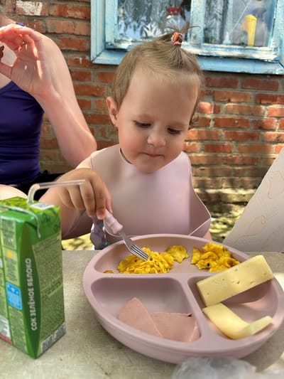In Komsomolskiy, Russia, a young child is seated outdoors at a table, eating scrambled eggs, cheese, and a slice of ham from a pink divided plate. The child is wearing a pink bib and is using a fork to eat. A green carton of juice with a straw is on the table next to the plate. In the background, a brick wall and a window with blue trim are visible.