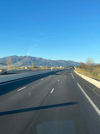 The image presents a forensic view of a multi-lane highway located within or near Le Boulou, France. The scene is captured under a clear, bright blue sky, with long shadows cast across the dark asphalt, suggesting either early morning or late afternoon.
The highway features at least two lanes of travel in the depicted direction, marked by white dashed lines, and is separated from opposing traffic by a concrete median barrier. A solid white line delineates the right shoulder, which is bordered by a concrete barrier and wild, dry vegetation including shrubs and grasses. Small white signposts are visible at intervals along the median barrier.
In the mid-ground to the left, a light-colored building or wall section is partially visible, along with several sparsely leaved deciduous trees. In the distance, a prominent range of mountains dominates the horizon. Several vehicles are observed traveling on the highway in both directions, appearing in the mid-ground and background. A faint, wispy contrail is present in the upper portion of the sky. No individuals are visible.