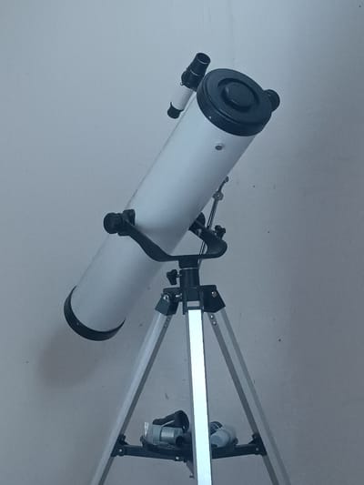 An image taken in Lisakovsk, Kazakhstan, shows a white telescope mounted on a silver tripod, positioned against a light-colored, textured wall. The telescope is cylindrical, with a black end cap on the far side and a smaller black eyepiece attached to the top near that end. The telescope is attached to a black mounting bracket, which is then secured to the tripod. The tripod has three legs made of silver metal. At the base of the tripod, a black tray-like accessory holds two cylindrical objects, one appearing to be a lens or eyepiece with a silver rim and a light blue tint, and another black object that could be a lens cap or cover. The lighting in the image is somewhat dim, casting a slight shadow from the telescope onto the wall. There are no people visible in the image.
