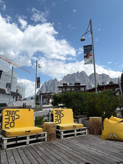 The image depicts an outdoor seating area in Cortina d'Ampezzo, Italy, with a backdrop of majestic Dolomite mountains. In the foreground, two bright yellow outdoor seating cushions with black lettering are placed on wooden pallets, serving as makeshift sofas. Next to them, a rough-hewn wooden log acts as a stool or side table. A large, yellow beanbag chair with a mountain logo is also visible. The wooden deck flooring contributes to a rustic, casual ambiance.
Behind the seating area, a lush green hedge provides a natural screen. In the mid-ground, several buildings with traditional alpine architecture are situated, some appearing to be hotels or residences. A lamppost with a banner featuring mountain imagery and the word "Cortina" suggests a connection to the town's identity.
Further in the background, a construction site is evident with a large yellow crane and scaffolding covering a building. The rugged peaks of the Dolomite mountains dominate the skyline, creating a dramatic and picturesque setting. The sky is a vibrant blue, dotted with fluffy white clouds, indicating fair weather. The overall scene conveys a sense of leisure and connection with nature, characteristic of a mountain resort town.