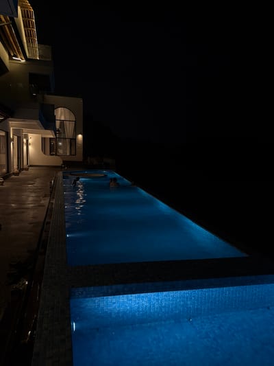 At night, a view from above shows a modern building complex with a long, narrow swimming pool in Jinhua, China. The building has geometric designs, including a rounded window. A wooden deck with railings lines one side. The pool is illuminated in shades of blue, with ripples and reflections on the water's surface. A textured, brick-like wall is visible to the right. The overall impression is one of contemporary design against the backdrop of the night sky. People are in the pool.