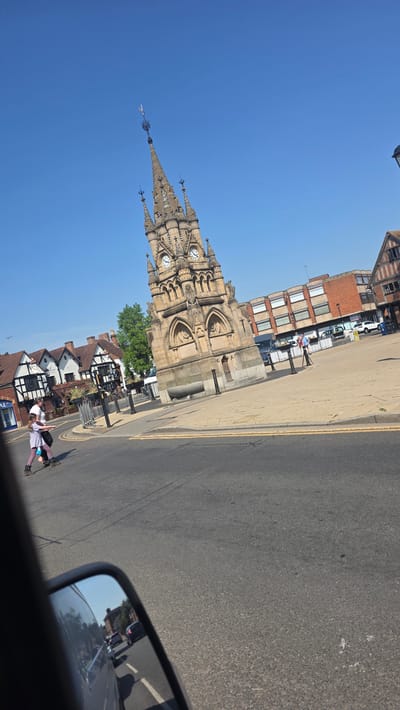 The image shows the High Cross monument in Stratford-on-Avon, United Kingdom. It is constructed of light brown stone. The monument has a clock on its upper section. Adjacent to it is a paved area, and next to that is a road surface. In the background, buildings stand. Two individuals are captured walking on the sidewalk near Tudor-style buildings with white walls and dark wooden beams. The sky above is clear and blue. A portion of a car's side mirror is visible in the foreground. The photograph is taken from a moving vehicle.