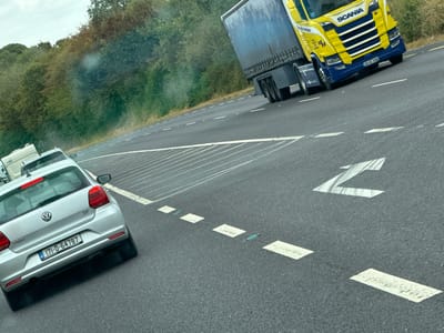 The image captures a dynamic scene on a highway, likely in Ireland given the license plates and road markings. A bright yellow Scania truck with a blue trailer is prominently featured, driving on the right lane of the asphalt road. The truck displays the license plate "201-KE-3398". In the left lane, a silver Volkswagen Polo hatchback is visible, with its rear end showing the license plate "171-D-64787". The car appears to be in motion. Beyond the vehicles, a dense line of green trees and foliage forms the backdrop, suggesting a rural or semi-rural setting. The sky is overcast, contributing to a muted lighting condition. Road markings, including white arrows and dashed lines, indicate lanes of travel. There are no visible people in the image. The context of Kilivory, Ireland, is not directly ascertainable from the visual elements alone, but the license plates are consistent with Irish registration.