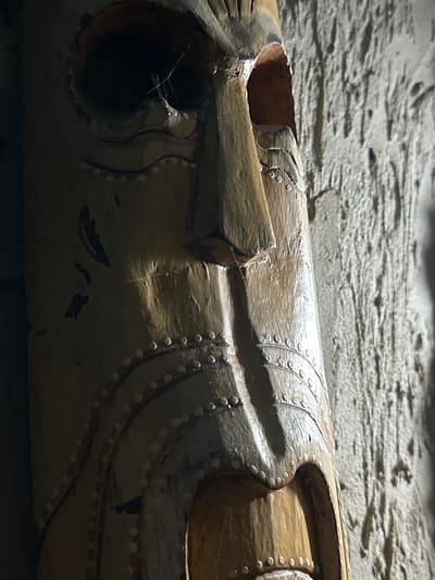 An extreme close-up, low-angle, vertical shot features a weathered, carved wooden mask against a textured, light-colored wall. The mask, oriented vertically and filling most of the frame, displays exaggerated facial features, including a prominent nose, large eye sockets (one dark, the other a gaping hole revealing an inner orange cavity), and a wide mouth. The surface of the mask is adorned with rows of small, circular indentations or beads, creating decorative patterns around the eyes, nose, and chin. Some areas of the mask show wear and tear, with dark smudges and a few strands of what appear to be spiderwebs visible within the left eye socket. The wall behind the mask is rough and uneven, with vertical striations and shadows. The lighting is dramatic, with strong highlights and deep shadows accentuating the mask's texture and form. The context provided places this image in Slov'yanoserbs'k, Ukraine, though no specific urban or environmental details beyond the wall and mask are discernible within the frame.