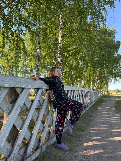 The image displays an outdoor scene in Ust'-Silayka, Russia, featuring an adult female leaning against a wooden fence. The woman, positioned on the left side of the frame, has long, light brown hair. She wears a dark plaid long-sleeved shirt and black pants with a repeating pattern of red chili peppers and white text. Her footwear consists of purple slipper-like shoes. Her left arm is resting on the top rail of the fence, and her left leg is bent with her foot placed on a lower section of the fence. Her head is turned to the right, and her gaze is directed away from the viewer.

The fence is constructed of weathered light gray wood, exhibiting some peeling paint. It features an upper section with diagonal lattice work and a lower section with vertical slats, extending from the foreground into the mid-ground along the left side of the image. Behind the fence and lining the path are multiple birch trees characterized by white bark and dense, bright green foliage. A clear blue sky is visible through the tree canopy.

A unpaved dirt path runs alongside the fence, extending from the mid-ground towards the background on the right. Long, distinct shadows from the fence slats and tree branches are cast across the path, indicating a low sun angle. In the far background on the right, more trees and open land are visible. Through a gap in the fence on the left, a glimpse of another fence and distant structures can be discerned. The environment suggests a rural or village setting during daylight hours.