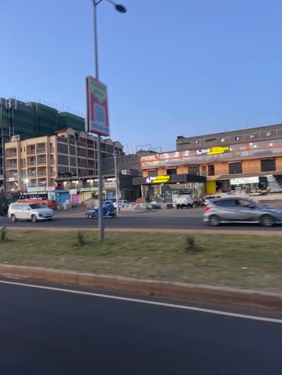 This image displays an urban street scene located in Biashara ward, Kenya, during either early morning or late afternoon. The foreground features a multi-lane asphalt road with a light brown concrete curb separating it from a green, sparsely vegetated median strip.

In the mid-ground, a white SUV and a blue motorized rickshaw (tuk-tuk) are observed moving from left to right, exhibiting motion blur. Simultaneously, a silver hatchback car is moving from right to left, also blurred by motion. A tall, dark street light pole with a vertically oriented, rectangular advertising sign, blurred but showing red and green elements, stands prominent on the left side of the median.

The background consists of various multi-story buildings. On the left, a residential building with several balconies appears to be under construction, evidenced by green scaffolding netting on its upper floors and exposed concrete block sections. To the center-right, a commercial building prominently features "Best Furniture" signage in yellow and purple lettering on both its ground and upper levels. Below this, various other smaller retail storefronts line the street. The sky above is clear and light blue. No specific individuals are clearly discernible within the frame.