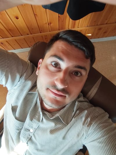 The image is a selfie taken by a person wearing a light-colored, pinstriped button-down shirt. The person has dark hair, brown eyes, and stubble on their chin and upper lip. The background of the image shows a wood-paneled ceiling and a brown upholstered chair. The lighting suggests the photo was taken indoors. There is no visible indication of the specific location within the city of Edineț, Moldova.
