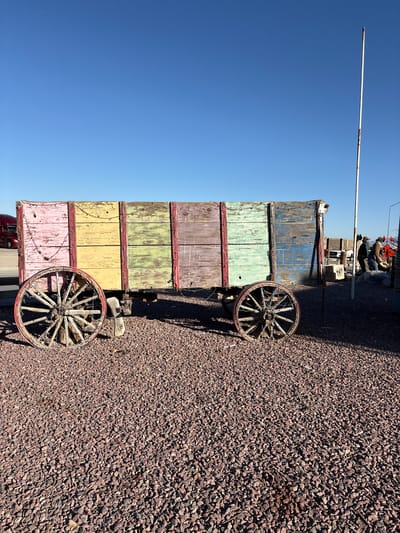 The image shows a vertical view of a weathered, multi-colored wagon with wooden wheels, parked on a gravel lot. The wagon is composed of several stacked sections, each painted a different faded color: blue on top, followed by green, yellow, and pink. The wood is visibly aged and worn, with peeling paint. Two large, spoked wooden wheels are attached to the wagon, one on each visible side. The wagon is positioned on a dark reddish-brown gravel surface.
In the background, out of focus, a few figures appear to be standing near some structures, possibly suggesting a market or outdoor display. A tall, thin white pole extends upwards into a clear, bright blue sky. The lighting suggests it is daytime, with a clear sky indicating fair weather. The overall scene evokes a sense of rustic charm and history, possibly hinting at an antique market, a historical reenactment site, or a decorative display. No discernible text is visible in the image.
