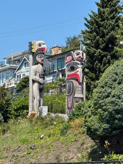 The image shows two totem poles in White Rock, Canada, on a sunny day. The totem poles are carved from wood and painted with intricate designs. The one on the left is taller and depicts a human-like figure holding a smaller figure. The one on the right has a more abstract design with bold colors. Both totem poles are surrounded by lush greenery, including trees and flowers. In the background, there are houses and a clear blue sky. The scene appears to be a public space, likely a park or a viewpoint, given the presence of the totem poles and the scenic setting. The lighting suggests it is daytime.