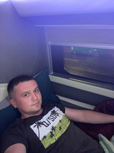 The image shows a man lying down, looking towards the camera. He is wearing a black t-shirt with a graphic design featuring palm trees and text. His head is resting on a dark blue pillow. To his right, a maroon blanket is visible. In the background, through a window, a green car is parked on what appears to be a street or parking lot. The scene is illuminated by a purple light source, likely from an LED strip, located above and to the left of the frame, casting a soft glow on the wall and ceiling. The overall environment suggests an interior setting, possibly a vehicle's sleeping cabin or a small room. There is no information in the image to suggest the location is in Ea Drăng, Vietnam.