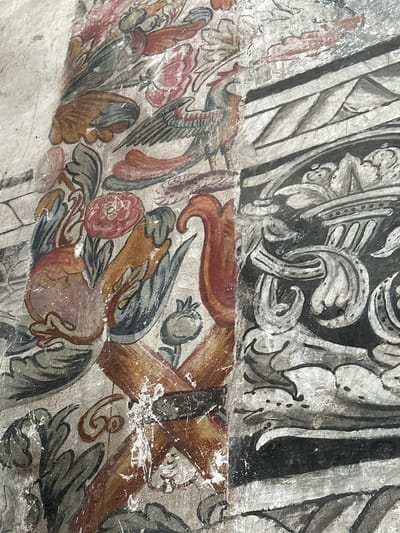 This is a detailed photograph of a fresco on a wall in Huejotzingo, Mexico, likely within the former Franciscan monastery. The fresco depicts a vibrant, stylized composition. On the left side, there is a section with botanical elements, including a reddish-pink flower resembling a rose, a round, reddish fruit, and a series of large, dark green, pointed leaves. A bird with intricate feathering in shades of blue, grey, and brown, with a red crest, is prominently featured. To the right of the botanical elements, the fresco transitions to a section with a more architectural and geometric design. This section features black and white patterns, including what appear to be mountain-like shapes within a banded structure, and elaborate scrolling white motifs against a dark background. The fresco exhibits signs of age, with visible cracks and areas where the paint has flaked away, revealing the underlying plaster. The colors, though somewhat faded, remain vivid, showcasing a rich palette of reds, blues, greens, browns, and black and white.