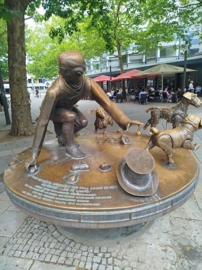 This media file captures a bronze sculpture on a circular base in an outdoor setting. The sculpture depicts a man crouching down, reaching out to a group of three dachshunds. The man is dressed in a jacket and boots, and his expression appears focused as he interacts with the dogs. One dog is looking up at him, another is sniffing the ground, and the third is walking away. There is also a top hat lying on the base near the dogs.

The sculpture is situated in a public space, possibly a plaza or street, with paved cobblestones around its base. In the background, there are trees with lush green leaves, suggesting it's daytime in a temperate season. Beyond the trees, buildings with windows are visible, and patrons are seated at tables under umbrellas, indicating a cafe or restaurant area. The overall atmosphere seems to be relaxed and leisurely.

Visible text on the base of the sculpture is in German. It appears to be a narrative or inscription related to the sculpture. Some legible phrases include "OFFENBACH BESUCHTE," "FRANKFURTER BÜRGERMANN," "DIE HUNDE LASSE SE LAAFE," and "WAR FESTGEFROREN." This suggests the artwork commemorates an event or story related to Offenbach and a citizen from Frankfurt. The time of day is likely daytime, and the weather appears pleasant, given the outdoor seating and foliage.
