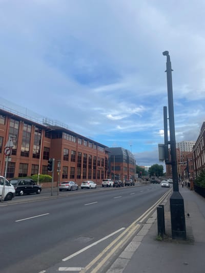 The image depicts a street scene in Reading, United Kingdom, likely on a cloudy day. The perspective is from a sidewalk, looking down a road lined with brick buildings. On the left, a large brick building with numerous windows dominates the foreground. A black taxi and a white van are parked on the side of the road in front of this building. Across the street, several cars are visible, including a silver Audi and a white Ford. Further down the road, traffic lights are red, and other vehicles are stopped. A tall black lamppost with attached signage stands prominently on the right side of the sidewalk. The sky is partly cloudy with a mix of blue and white. The overall impression is an urban street scene with typical city infrastructure and traffic.