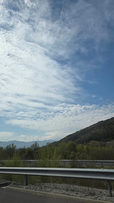 The image shows a view in Jeleznitsa, Bulgaria. The sky is blue with extensive altocumulus clouds. A wooded hillside is visible to the right, and a section of highway infrastructure, including a metal guardrail and asphalt road surface, is in the foreground.