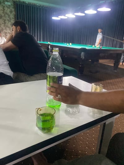 The image depicts an indoor scene, likely a billiard hall in Боғистон, Tajikistan. In the foreground, a white table is prominent, holding a plastic bottle of green liquid and a glass also filled with the same green liquid. A person's arm and hand are visible, reaching towards the bottle. In the background, a pool table with several balls is set up under overhead lights. Two individuals are partially visible near the pool table; one, wearing a patterned shirt, stands by the table, while another, wearing a black t-shirt, is seated and leaning forward. The environment features dark curtains along the wall and a patterned carpet on the floor.