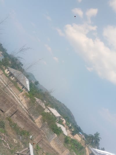 The image depicts an elevated view of a residential area situated on a hillside in Awo Ekiti, Nigeria, captured with a diagonal orientation. The upper-right portion of the frame shows a light blue sky with scattered white cumulus clouds. A faint, small red circular mark is present in the clear sky region, near the top-center of the rotated image.
The terrain slopes downwards from the left foreground towards the right background, indicating a settlement built into a hill. Numerous single-story structures, primarily houses, are visible across the mid-ground and background. Many buildings feature metallic corrugated gable roofs, while others appear to have flat concrete roofs or are in various stages of construction, displaying exposed concrete block walls. Concrete block fences and metal gates define property boundaries between structures.
Sparse vegetation, including grass and low-lying shrubs, covers the immediate foreground and areas around the buildings. Denser foliage, comprising various trees, tall bamboo-like stalks, and palm trees, is distributed throughout the scene, particularly on the hillside beyond the houses and amidst the built-up areas. No individuals, animals, or specific actions are discernible within the frame. The overall impression is a semi-urban or rural settlement integrated into a natural, hilly landscape under daylight conditions.