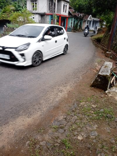A white car is driving on a paved road. The car is heading away from the camera.  It is a small hatchback car, with black wheels and tinted windows. The car is driving on a narrow road with a dirt shoulder. The road is located in Kecamatan Cisewu, Indonesia, at coordinates -7.383, 107.504. There are two motorcycles parked on the right side of the road. The motorcycles are both black and have a rider on them.  The motorcycles are driving on a paved road, which is curving to the right, and there are buildings on the left and right sides of the road. In the foreground, there is a patch of grass and some dirt.