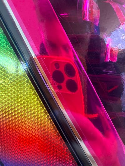 This image is a close-up, abstract shot of a phone case with a holographic, iridescent design. The primary colors are vibrant pinks, purples, yellows, and reds, creating a rainbow effect. The surface is highly reflective, showing distorted reflections of other objects, including what appears to be a hand holding the phone and potentially the environment around it.

In the foreground, a portion of a phone is visible, featuring a distinctive camera array with four circular lenses arranged in a square pattern. The phone itself appears to be red or a similar warm hue, with its reflection prominently displayed on the holographic material.

The holographic surface is textured, with a diamond-like pattern visible in the yellow and red sections. The material seems to be a film or coating applied to the phone case, creating a shimmering, futuristic aesthetic. The lighting is bright and likely artificial, enhancing the reflectivity and saturated colors of the phone case. There are no discernible signs of a specific time of day or weather conditions, and the setting is implied to be indoors or in a controlled environment where the phone is being showcased or used. No text is visible in the image. The overall impression is one of modern technology with a playful and eye-catching design.