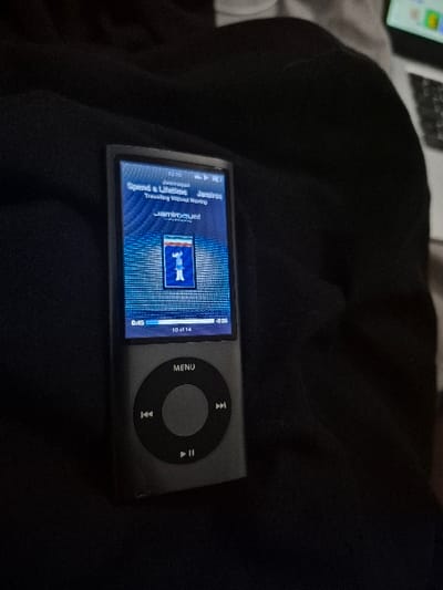 The image shows a black iPod classic resting on black fabric. The screen displays the song "Spend a Lifetime" by "Jamiroquai" playing from the album "Travelling Without Moving." The time displayed is 10:01:14, and the track is 3:25 long. The iPod is in portrait mode. The location is 51.882 latitude and 0.144 longitude, which is in a residential area in East Hertfordshire, United Kingdom. The scene is dimly lit, and the fabric appears to be a piece of clothing.