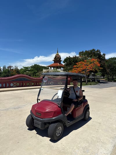 A man is sitting in a red golf cart on a concrete surface, with a building with ornate orange roofs and golden spire visible in the background. The number "221" is visible on the golf cart. A red fence with a dragon design is visible next to the cart. Trees and a clear blue sky are also visible. The location is Bang Pu Subdistrict Municipality, Thailand.