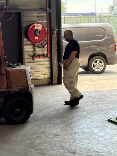 The image shows a man walking in front of a closed corrugated metal garage door. A red fire hose reel is mounted on the wall to the left of the door. A grey van is parked to the right of the garage door. A forklift is partially visible in the foreground to the left of the man. The man is wearing a dark t-shirt and light-colored work pants. He is bald and has a slight smile on his face. The scene appears to be an industrial or commercial setting. There is no identifiable information that places the image in the city of Mol, Belgium.