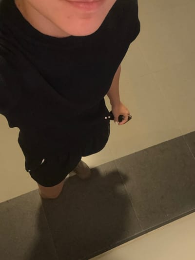 A high-angle, downward perspective captures a partial view of an individual standing indoors. The subject, visible from the mid-chest to the feet, wears a black short-sleeved top or dress and black shorts or skirt. A light-colored shoe is on the left foot. The right hand is visible, holding a black smartphone accessorized with white-tipped elements, and the fingers display white nail polish. A subtle smile is present on the visible lower lip and chin. A small, light-colored object is partially visible protruding from the left side of the black shorts/skirt. The environment consists of a tiled floor divided into two sections: a lighter beige/off-white section in the upper half and a darker gray rectangular-tiled section in the lower half, separated by a distinct horizontal line. Artificial lighting casts a clear shadow of the person onto the dark gray floor. The specific location within the city of Santa Catarina, Mexico, is not discernible from the visual evidence.