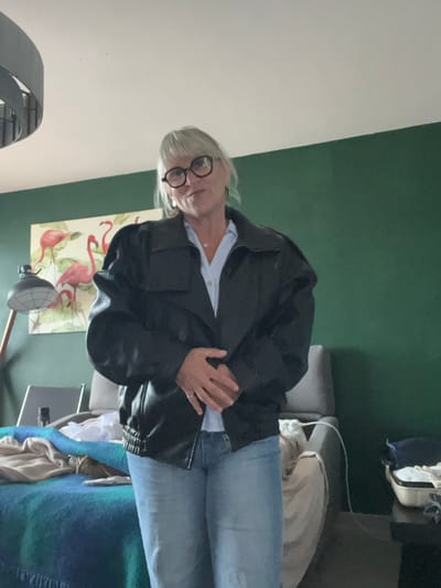 A person wearing glasses, a light-blue button-down shirt, a black leather jacket, and light-blue jeans stands in front of a wall that is painted green in the top half and white in the bottom half. On the lower half of the green wall, a painting of pink flamingos on a beige background is partially visible. A gray standing fan is below the painting. A gray couch covered with a blue and green blanket is on the right side of the image. In the background, a white bag sits on top of the couch. There is not sufficient information to determine the location in Palavas-les-Flots, France.