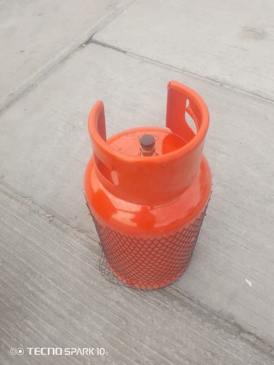 The media file is a photograph of an orange gas cylinder resting on a concrete surface. The cylinder is the primary object, with a black mesh netting around its lower half. It has a cap on top and handles on its upper section. The concrete surface appears weathered, with visible cracks and lines indicating separate slabs. The lighting suggests it is daytime, with no specific weather conditions evident. The image is taken from a slightly elevated angle, looking down at the cylinder. The background is out of focus, but appears to be more of the same concrete ground. Visible text at the bottom of the image reads "TECNO SPARK 10c". The scene is simple and focuses on the gas cylinder.