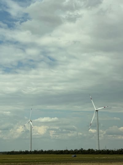 An outdoor, low-angle, full shot captures two white wind turbines in a field under a cloudy sky. The turbine on the left is closer to the viewer, while the turbine on the right is slightly further away. The turbines have three blades, each with red accents on the tips. In the foreground, a green field is visible with a dark-colored SUV parked on the far left. The background features a dense line of green trees, behind which the turbines stand. The sky is filled with scattered clouds of varying gray and white tones. The image appears to be taken from a vehicle traveling through a rural area, possibly on the outskirts of Mayak, Russia.