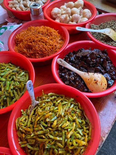 This is a high-angle, close-up shot of several red bowls filled with various food items, likely prepared to be served as side dishes or condiments. The bowls are arranged on a wooden surface, suggesting an outdoor market stall or a home kitchen setting in Chongzhou City, China.

In the top left bowl, there is a pile of finely ground spices or seeds, with a white and yellow spatula resting within. Adjacent to it, to the right, is a bowl filled with dark, seemingly pickled or preserved food pieces, a wooden spoon with red chili flakes scattered on it is placed inside. To the right of this, a large bowl is filled with thin, green, julienned vegetables, possibly pickled or stir-fried, with a spoon in it.

Below the spices, a bowl contains whole, peeled garlic cloves. Next to the garlic, to the right, is a bowl filled with thin, orange strands, resembling shredded vegetables or noodles, possibly pickled. Further to the right and below the green vegetables is another bowl containing similar green, julienned vegetables.

There's a glimpse of a metal container with a silver top, possibly a grinder or a dispenser, to the left of the orange strands. White plastic bags with some green print are visible at the bottom of the frame, and some Chinese characters are visible on one of the bags. The lighting suggests it's daytime. The overall impression is of a colorful and diverse selection of food preparations, typical of an Asian culinary context.