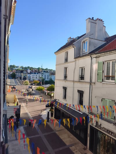 The image shows a street scene in Lagny-sur-Marne, France, on a clear day. A pedestrian walkway is decorated with colorful pennant banners strung across the street. To the right, a building with a white facade and green shutters is visible, with a shop sign that reads "JM. COIFFEUR". In the background, there are more buildings and a hill covered with trees and houses. Several parked bicycles are also visible.