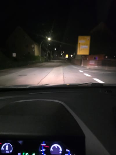 This detailed description pertains to a nighttime scene captured from inside a vehicle on a road in Waldmünchen, Germany. The perspective is looking forward through the windshield, which has visible dust and debris.
In the immediate foreground, the top portion of a car's dashboard and instrument cluster is visible, illuminated by internal lights. The visible gauges include a digital display showing "15c" for external temperature.
The car's headlights brightly illuminate the asphalt road directly ahead, revealing some minor imperfections and a centerline or seam. On the right side of the road, a series of dashed white lines demarcates a lane or the edge of the roadway.
On the left side of the road, a multi-story building with a dark, pitched roof is partially visible against the black night sky. It appears to be a residential structure, with one window on the lower floor casting a faint yellow light. A streetlight, partially obscured, is positioned further down the road on the left, emitting a bright glow.
On the right side of the road, further in the distance, a prominent yellow rectangular German directional sign is visible, although slightly blurry. The sign clearly indicates "Waldmünchen" as a destination, likely straight ahead, and another destination, "Rötz," with an arrow pointing to the right. Beyond this sign, a reddish-brown building, possibly commercial, with dimly lit sections, stands along the road. Another streetlight is visible further down the road on the right.
The scene is located on **Böhmerstraße in Waldmünchen, Germany**, specifically looking south along the road in the vicinity of the intersection with W. Jäger-Straße, where a right turn would lead towards Rötz.