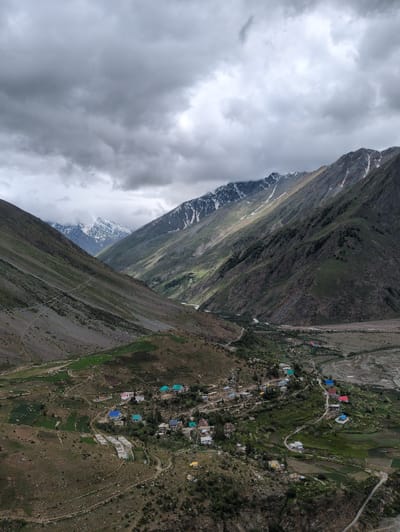 An aerial drone shot captures a village in the Darcha region of India, nestled within a valley of imposing, rugged mountains. The sky above is heavily overcast with dark, dramatic clouds, suggesting impending or recent rain. The slopes of the mountains are a mix of rocky terrain and patches of green vegetation, with some areas showing signs of cultivation. A winding dirt road snakes through the landscape, connecting the scattered cluster of buildings that form the village. The houses are of various colors, with some featuring brightly colored roofs in shades of blue, red, and turquoise. A river or stream is visible in the distance, carving its way through the valley floor. The overall scene is one of remote, natural beauty, characteristic of mountainous regions.