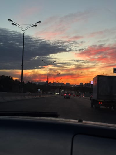 The image depicts a view from a vehicle traveling on a multi-lane highway at sunset. The sky is filled with vibrant colors, including hues of orange, pink, and red, with streaks of darker gray clouds interspersed. Several streetlights, characteristic of urban roadways, line the highway. In the distance, city buildings are visible on the horizon. Traffic is present, with cars and a large truck on the highway. The foreground shows the interior of the vehicle from which the photograph was taken, specifically the dashboard and windshield area. The overall impression is a typical urban commute during a scenic sunset.