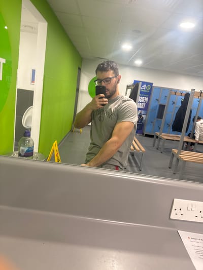 The image is a selfie taken in a locker room, likely in a gym or sports facility. The location is Sheffield, United Kingdom, based on text visible in the image. The person taking the photo is a man with short dark hair, a beard, and glasses. He is wearing a grey t-shirt and appears to be standing in front of a mirror. In the background, there are rows of blue lockers and wooden benches. A wet floor sign is visible. A bottle of water is on the sink. The room has green and white walls.