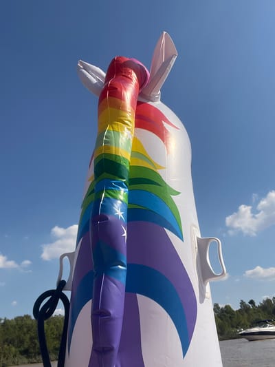 The image shows a close-up of an inflatable unicorn float against a blue sky with scattered clouds. The unicorn is white with rainbow-colored sections. Its tail is a rainbow-striped tube with white stars. The photo appears to be taken outdoors on a sunny day. The location in San Fernando, Argentina is not visible in this image.