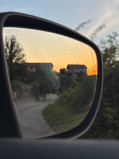 The image is a reflection in a car's side mirror, showing a rural scene in Svinjišta, Montenegro at sunset. The mirror reflects two houses nestled amongst trees on a hill. A dirt road curves up towards the houses, with green foliage and trees lining the sides. The sky is a warm orange and yellow, characteristic of a sunset, with some wispy clouds visible. The car's interior is partially visible in the foreground, framing the reflection.