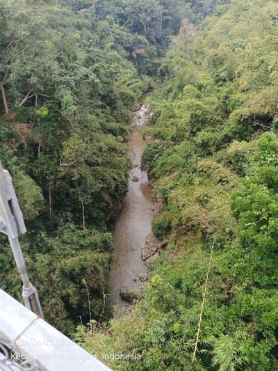 An overhead view captures a winding river flowing through a lush, densely forested gorge. The river, appearing muddy, is bordered by rocky banks and dense green foliage composed of trees and bamboo. The scene is a natural landscape, with no people or animals visible. The image is dated 2026/06/23 at 12:23 and the location is identified as Kecamatan Kuningan, Indonesia. A metal cable and support structure are visible in the lower left portion of the frame, suggesting the viewpoint is from a bridge or elevated platform.