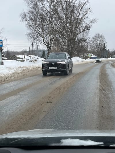 The scene depicts a grey winter day on a two-lane paved road in Tatishchevo, Russia. A dark grey or black Changan SUV, bearing Russian license plate X745MB 799, drives towards the viewer in the left lane. Its daytime running lights are illuminated. The road surface is wet, showing streaks of melted snow, mud, and slush, with compacted snow and dirt visible on the shoulders. Bare deciduous trees line both sides of the road, and in the background to the left, several single-story buildings are visible amidst snow-covered ground. A blue bus stop sign is present on the left side of the road. Further down the road on the right, a white or silver sedan is visible driving away from the viewer, near a pedestrian crossing sign. The image appears to be taken from inside another vehicle, with the top of its dashboard visible at the bottom of the frame. No individuals are explicitly visible.
