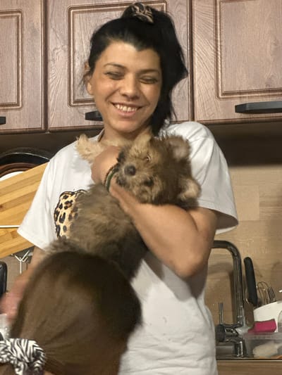 A woman with dark hair pulled into a bun holds a small, fluffy brown dog in her arms. She is wearing a white t-shirt with a leopard print design on the chest and a beaded bracelet on her wrist. The background appears to be a kitchen, with wooden cabinets and a sink visible. The scene suggests a casual, domestic setting. The location is in the city of Ruska Bela, Bulgaria.