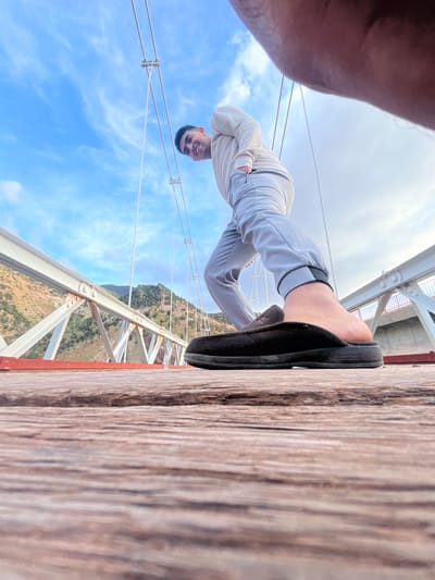 A young man stands on a wooden structure, possibly a bridge, with a mountainous landscape and blue sky with scattered clouds visible in the background. The bridge appears to have white metal supports and cables. The man is wearing a long-sleeved white shirt, grey pants, and black sandals. The photograph is taken from a low angle, with part of the photographer's arm visible in the bottom right corner. The location is likely Tabaroucht, Morocco, but not clearly visible in the image.