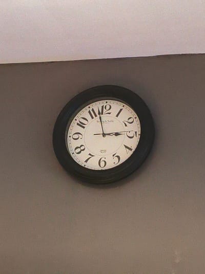 The media file shows a close-up of a round wall clock with a black frame. The clock face is white with black Arabic numerals. The hands of the clock are also black. The hour hand is pointing slightly past the 10, and the minute hand is pointing at the 5. Therefore, the time shown on the clock is approximately 10:25. There is some faint text on the clock face, possibly a brand name or logo. The clock is mounted on a plain, taupe-colored wall, and the top edge of the frame or ceiling is visible at the very top of the image. The lighting appears to be ambient, suggesting an indoor setting. There are no people or other objects in the frame besides the clock. The overall impression is that of a functional, everyday object in a simple, unadorned environment. The location context of Cavite City, Philippines, does not provide specific visual cues within the image itself, but it establishes the geographical setting.