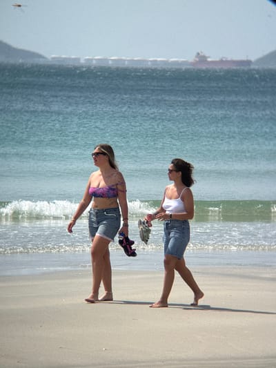 Two women are walking on a sandy beach with the ocean in the background. The woman on the left is wearing a strapless pink and purple patterned bikini top and denim shorts. She has sunglasses on and is carrying a pair of dark flip-flops and a can in her right hand. The woman on the right is wearing a white spaghetti strap tank top and denim shorts. She is also wearing sunglasses and carrying a pair of flip-flops in her left hand. The water is a clear blue-green and there are small waves breaking on the shore. In the distance, a long cargo ship is visible on the horizon. The scene appears to be a sunny day. This location is not in Dong Ninh Hoa, Vietnam.