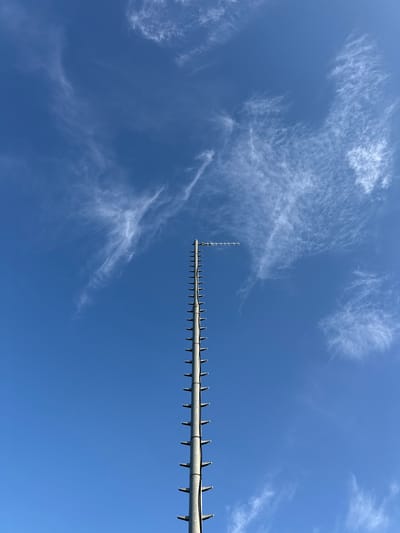A tall, slender metal pole extends upward into a bright blue sky, its surface marked by regularly spaced horizontal protrusions resembling rungs or fixtures. At the very top of the pole, a horizontal antenna array is visible. Wispy, white cirrus clouds are scattered across the sky, creating a delicate, streaky pattern against the deep blue. The perspective is from below, looking up at the pole and sky. The image appears to be taken outdoors in a location that allows for a clear view of the sky and a tall structure, potentially in a less densely populated area given the open sky and lack of surrounding urban features. The location is the village of Nučice, Czechia.