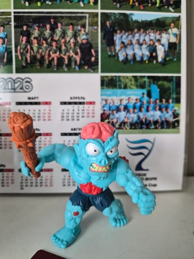 A blue zombie toy with a pink brain, wearing dark blue shorts and a red chest piece, stands on a light-colored surface. The toy is holding a brown mace-like object in its right hand. Behind the toy is a calendar for the year 2026. The calendar has sections for March and April visible. The background also shows images of sports teams, possibly soccer teams, with players in uniforms. One image shows a team in green uniforms, another in white and blue, and a third group of people in blue uniforms. The overall setting appears to be indoors, with the calendar and photographs suggesting a desk or wall display.
