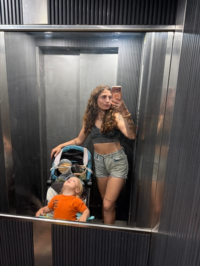 An interior shot, likely a selfie taken in a mirror or reflective wall, features a woman and a child. The woman, positioned on the left, has long, wavy brown hair and visible tattoos on her left arm and left thigh. She wears a patterned crop top and light-colored denim shorts, holding a smartphone with an orange case in her right hand to capture the image. Her expression is neutral as she looks towards the camera.
Beside her, on the right, is a young child with light blonde hair, dressed in an orange t-shirt with a "222" pattern. The child is seated or lying in a blue and white stroller, looking upwards. A pacifier is visible in the child's left hand, and clear plastic containers, possibly holding food, are placed in the stroller.
The background, reflected in the surface, consists of dark, horizontally ribbed or corrugated walls, suggesting an enclosed space such as an elevator or a modern corridor. The overall setting is inside a building in Khalde, Lebanon. The lighting is artificial, suitable for an indoor environment.