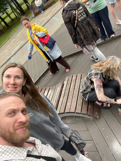 The image shows a group of people in an outdoor setting, possibly a park or a recreational area, as indicated by the wooden decking, benches, and trees visible in the background. The people are in casual attire, and their poses suggest a relaxed and informal gathering. One person is sitting on a bench, wearing a plaid shirt and dark pants, with visible tattoos on their arm. Another person is standing and holding a blue object. Two people are in the foreground, appearing to be taking a selfie. One of them is a woman with long brown hair and a light-colored hoodie, looking at the camera with a smile. The other is a man with a beard and a checkered shirt, also looking towards the camera. There is a colorful jacket with a backpack in the middle ground. A bicycle is partially visible.

The time of day appears to be daytime, with natural lighting. The weather seems mild, and the overall mood is lighthearted and friendly. The setting might be a place for leisure activities.

Visible text includes "NEW YORK LONDON MUNICH BERLIN", "SEWENCHO", and what appears to be Cyrillic characters "МИЛ", on the clothing of some individuals. The location context of Sosnoviy Bor, Russia, suggests a specific geographical area for this scene.