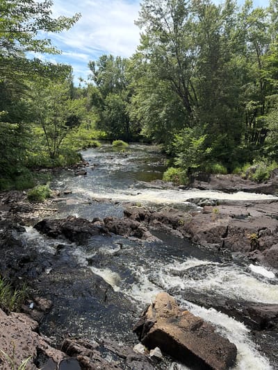 The image shows a rocky river flowing through a lush, green forest. The water is dark and turbulent, with white foam indicating rapids. Large, dark rocks are scattered along the riverbed and banks. The trees are dense and tall, with sunlight filtering through the leaves. The sky is blue with scattered white clouds. The scene appears to be a natural landscape, likely in a wilderness area.