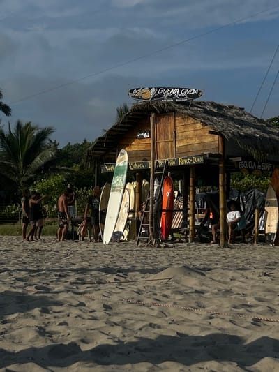 The image shows an outdoor scene in Montañita, Ecuador, located at the coordinates -1.825 latitude and -80.754 longitude. The scene features a wooden structure covered with a thatched roof and a sign overhead reading "Buena Onda Surf School, Montañita, Ecuador". There are multiple surfboards leaning against the structure, and a few people are standing and sitting in front of it. Several of them are wearing swimsuits, and some are wearing sunglasses. The people are interacting with each other, and some are talking and gesturing. The ground is covered in sand, indicating a beach setting. In the background, there are palm trees and other vegetation. The sky is mostly clear, with a few clouds in the distance. The overall ambiance is relaxed and casual, suggesting an environment conducive to water sports and beach activities.