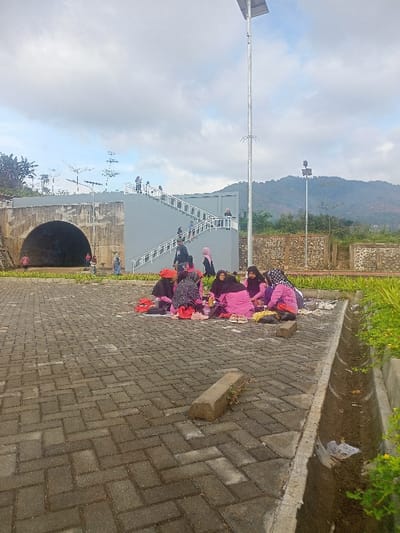 The image depicts an outdoor scene in what appears to be a public or recreational area in Kecamatan Pangalengan, Indonesia, under an overcast sky. In the foreground, a group of approximately eight women are seated on a paved surface made of gray interlocking bricks. Many of the women wear headscarves (hijabs) in various colors including pink, purple, and black, and they appear to be picnicking or socializing, with small red bags and other items scattered around them. A concrete block and a drainage ditch are visible on the right side of the foreground.
In the midground, a large concrete structure with a distinctive gray color features a prominent arched tunnel entrance on the left. Adjacent to it on the right is a multi-level staircase with white railings, where several people are seen walking up and down. Above the tunnel structure, there are decorative elements resembling abstract trees or light fixtures. A very tall light pole with a solar panel on its top stands in the center-right of the frame, with another smaller pole visible further in the background. Behind the group of women and extending to the right, there's a textured stone wall.
The background reveals distant, undulating green hills and mountains under a cloudy sky. The lighting is soft and diffused, indicating a daytime setting, possibly morning or late afternoon, without direct harsh sunlight. The overall atmosphere seems relaxed and casual, suggesting a leisurely gathering. No discernible text is visible in the image.